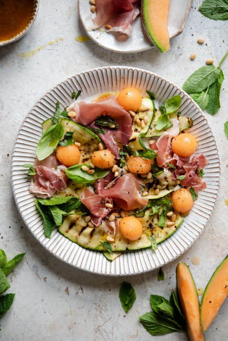 A colorful bowl of grilled zucchini salad topped with melon and prosciutto.