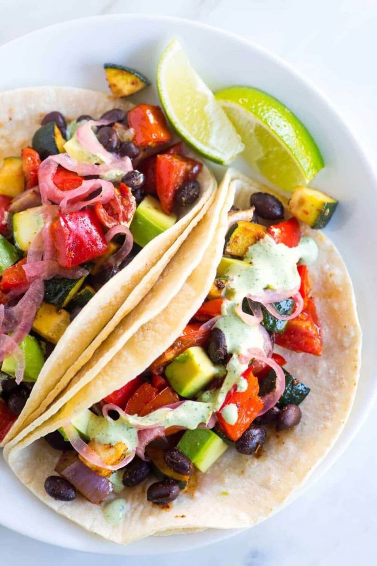 A closeup of a plate of colorful roasted veggie tacos topped with creamy cilantro sauce.