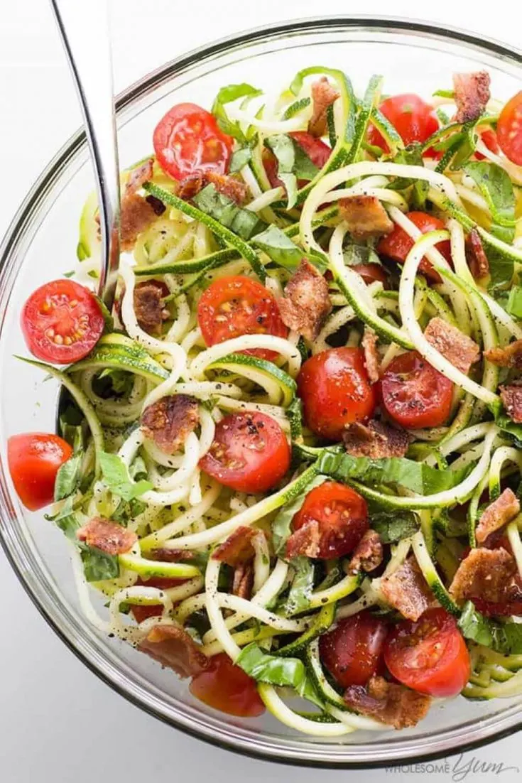 A large bowl of zucchini noodle salad topped with bacon and tomatoes.