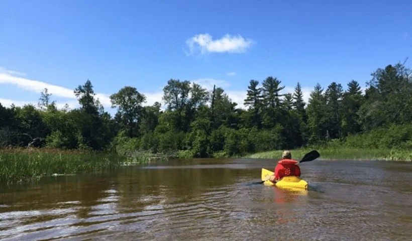 A man kayaking down the Au Train River with forest and grass on either side of the water.